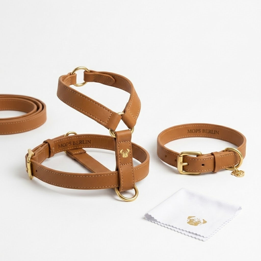 Ein Set aus handgefertigtem Hundegeschirr und passendem Halsband von MOPS BERLIN, aus cognacfarbenem veganem Leder mit messingfarbenen Beschlägen, ordentlich präsentiert auf weißem Hintergrund neben einem gebrandeten Reinigungstuch.