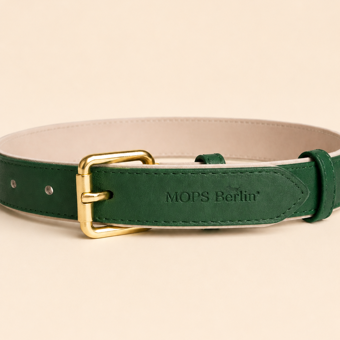 Nahaufnahme der mattgoldenen Schnalle und der eingeprägten Markenprägung MOPS Berlin auf dem dunkelgrünen veganen Lederhalsband Goldbär vor beige-farbenem Hintergrund.