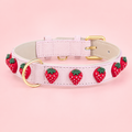Quadratisches Produktfoto des Mops Berlin Hundehalsbands Erdbeersommer aus rosafarbenem veganem Leder, verziert mit roten Erdbeer-Applikationen sowie goldener Schnalle und D-Ring vor rosa Hintergrund