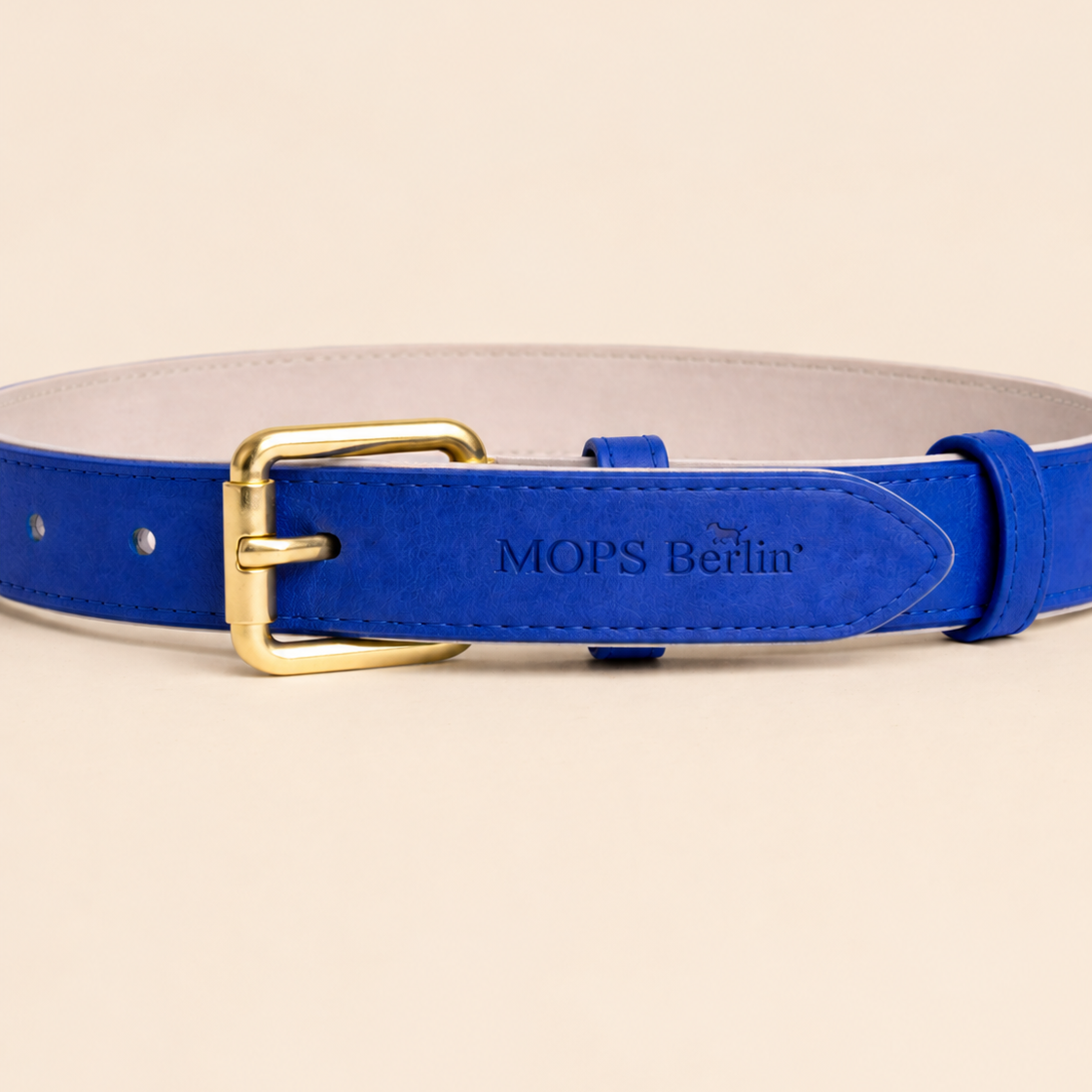 Nahaufnahme vom Schnallenende des Mops Berlin Hundehalsbands Sternennacht aus blauem veganem Leder mit eingeprägtem Logo und mattgoldener Metallschließe vor beige-cremefarbenem Hintergrund